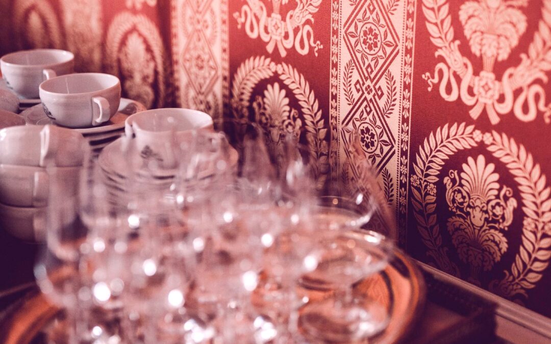 Gläser und gestapelte weiße Tassen vor einem dekorativen Wandbehang mit floralen Mustern