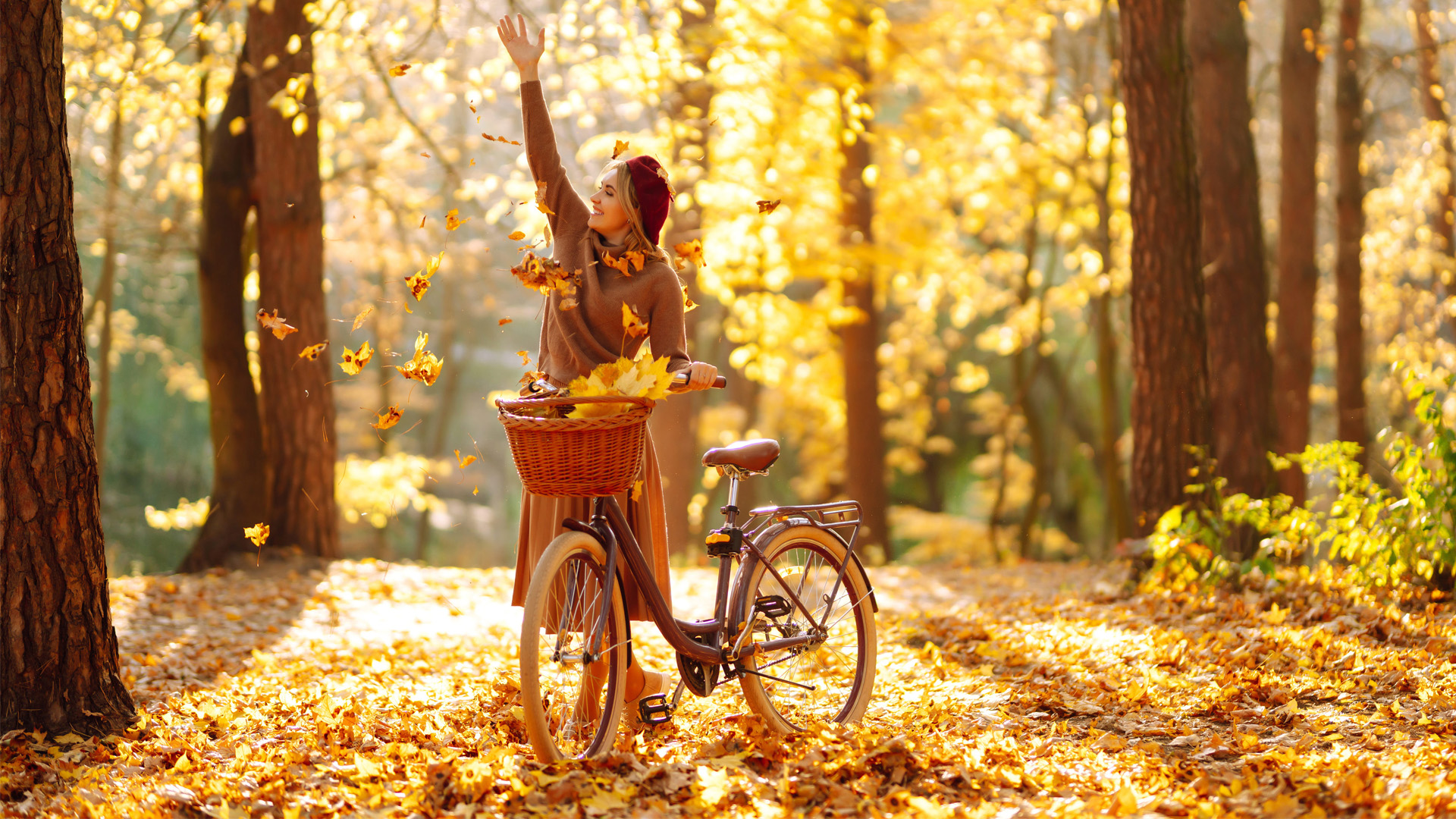 Herbstspecial Frau in braunem Kleid wirft bunte Herbstblätter in die Luft neben einem Fahrrad auf einem von Bäumen gesäumten Waldweg.