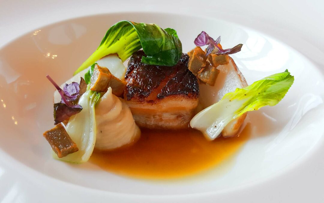 Gebratener Fleischwürfel in brauner Sauce, umgeben von kleinen grünen Blättern, weißem Gemüse und braunen Würfeln.